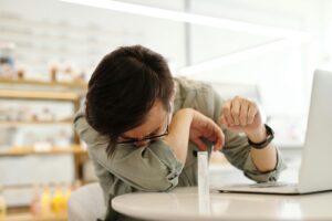 An adult man sneezes into his arm near a laptop in a cafe, illustrating illness prevention.